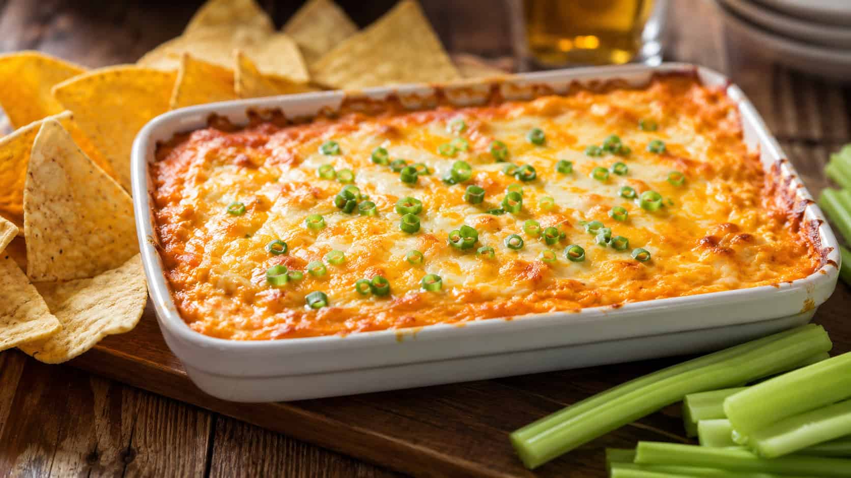 a bubbling baking dish of buffalo chicken dip in a wide horizontal shot, golden melted cheese on top and sliced green onions, surrounded by tortilla chips and celery sticks, rustic table, warm natural light, shallow depth of field, landscape orientation
