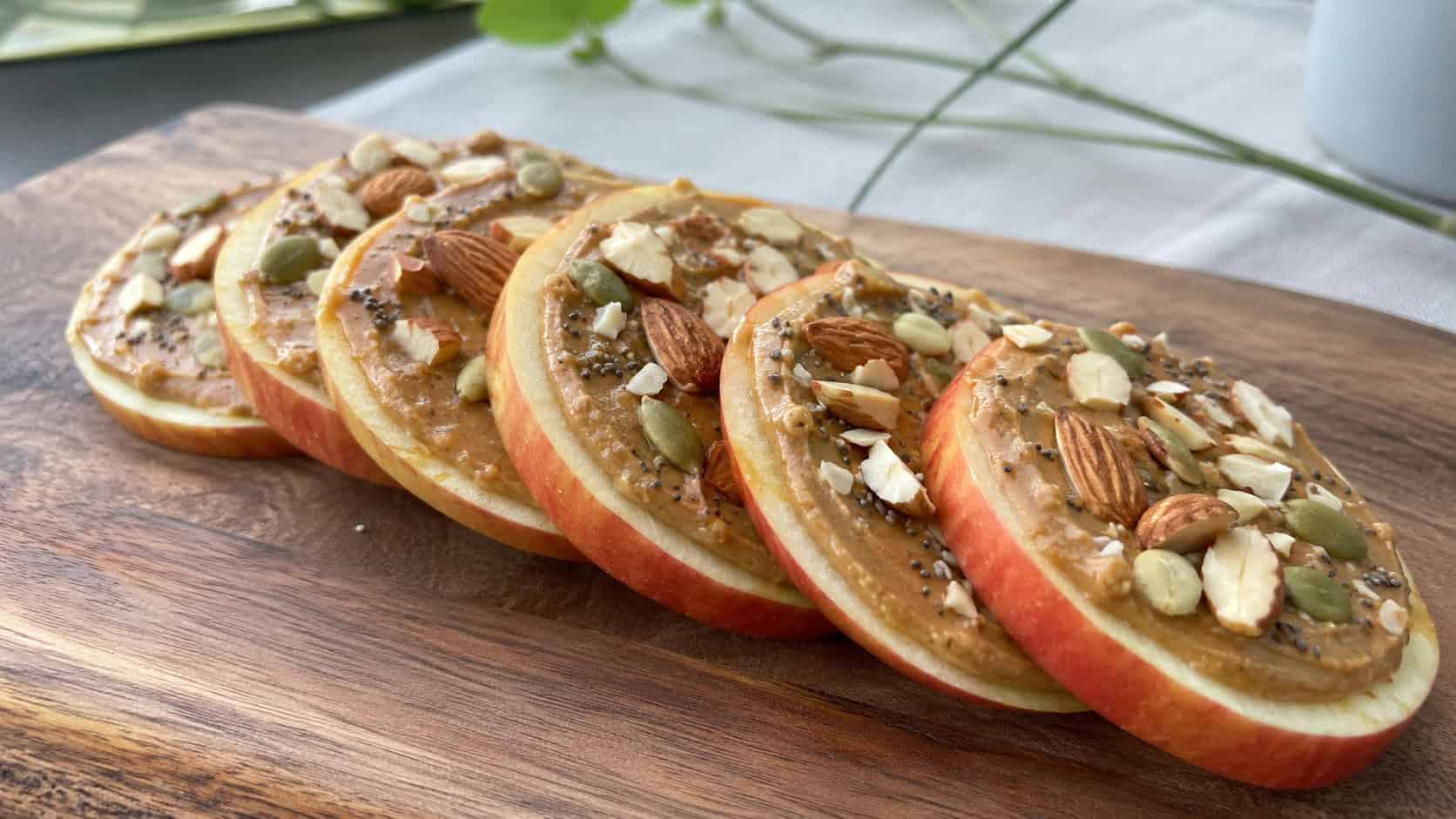 almond butter apple slices in a wide horizontal shot, apple rounds spread with almond butter and topped with seeds and almonds, fanned out on a wooden board, bright natural light, shallow depth of field, landscape orientation