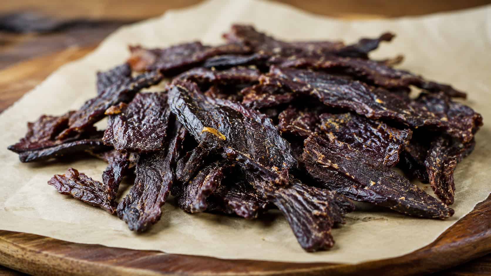 homemade beef jerky bites in a wide horizontal shot, dark chewy strips of marinated beef piled on parchment paper, on a wooden board, warm natural light, shallow depth of field, landscape orientation