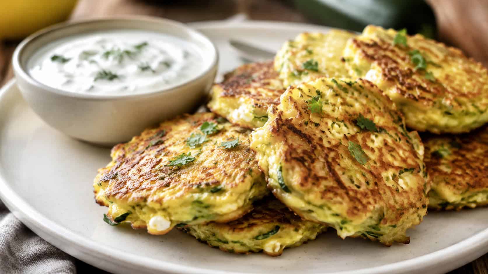 zucchini feta fritters in a wide horizontal shot, golden crispy fritters flecked with green zucchini stacked on a plate with a yogurt dip, warm natural light, shallow depth of field, landscape orientation