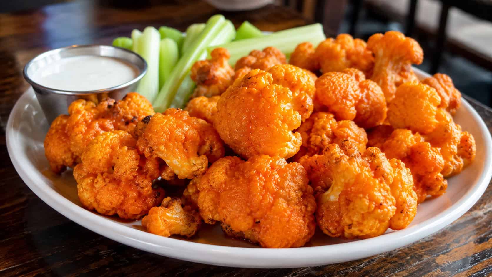 buffalo cauliflower bites in a wide horizontal shot, crispy orange-coated cauliflower florets on a plate with celery and a dipping cup, warm natural light, shallow depth of field, landscape orientation