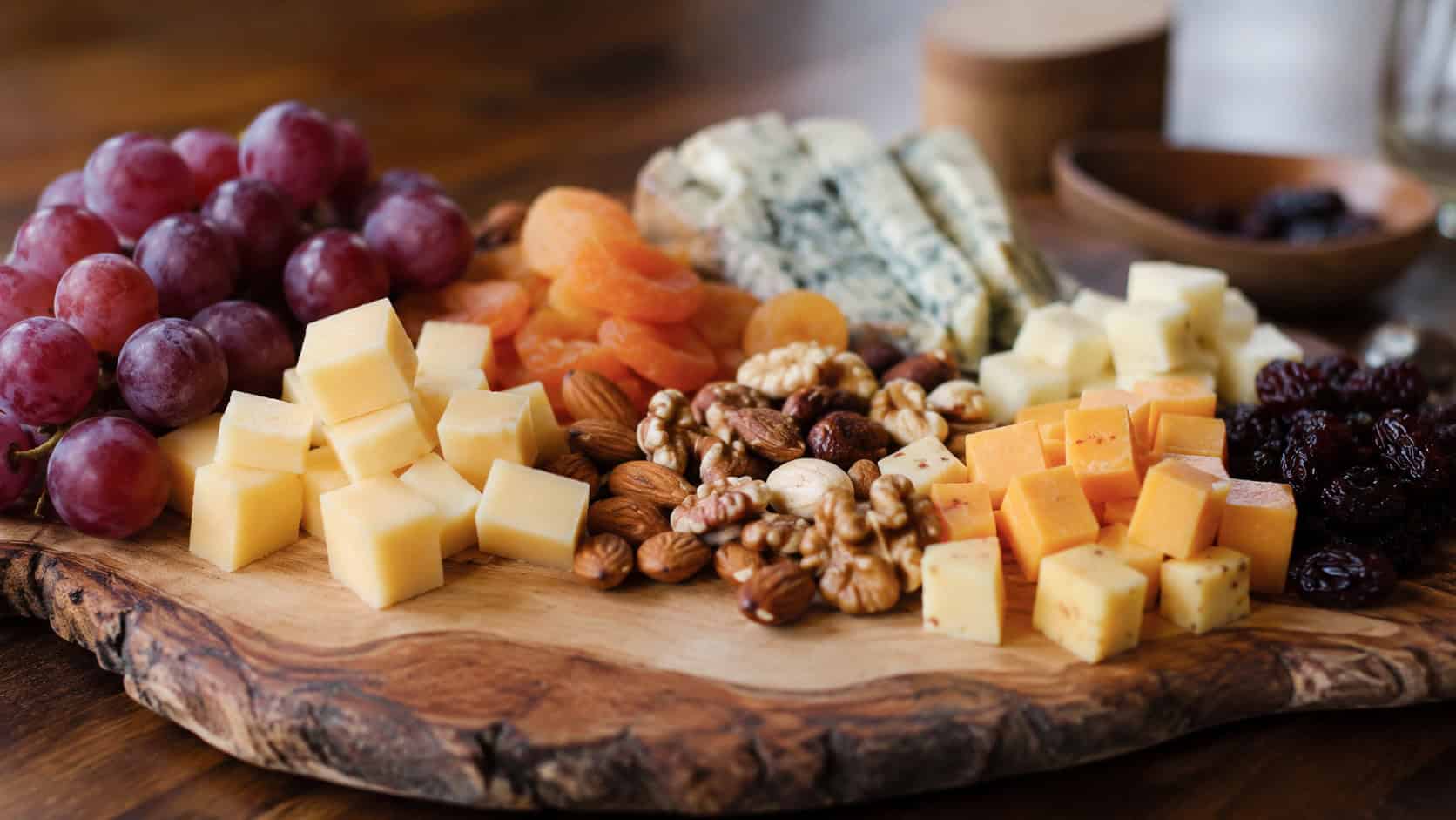 a cheese and nut platter in a wide horizontal shot, an assortment of cubed cheeses, mixed nuts, grapes, and dried fruit arranged on a wooden board, warm natural light, shallow depth of field, landscape orientation