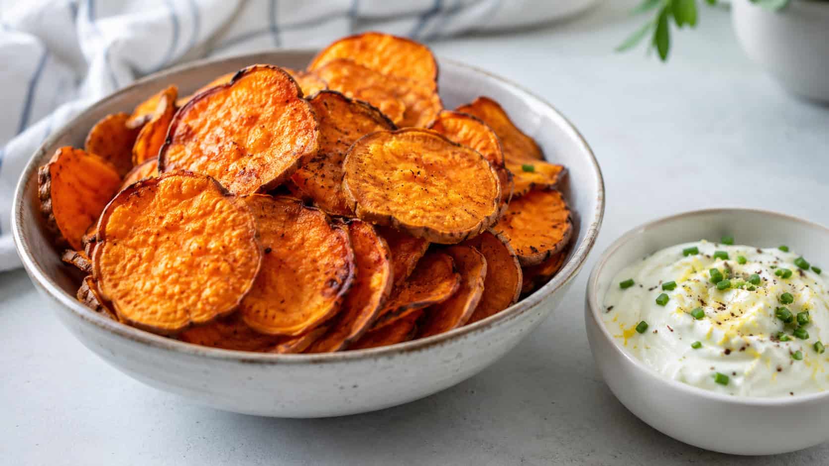 baked sweet potato chips in a wide horizontal shot, crispy orange chip rounds piled in a bowl beside a creamy yogurt dip, on a light surface, bright natural light, shallow depth of field, landscape orientation