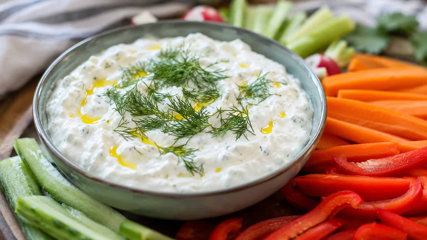 Greek yogurt dip in a wide horizontal shot, a thick white dip topped with fresh dill and a drizzle of olive oil in a bowl, surrounded by vegetable sticks, bright natural light, shallow depth of field, landscape orientation