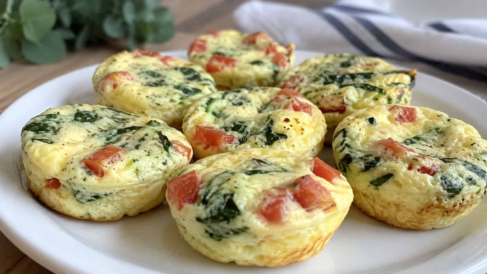 egg white frittata bites in a wide horizontal shot, pale fluffy egg muffins flecked with red tomato and green spinach on a white plate, bright natural light, shallow depth of field, landscape orientation