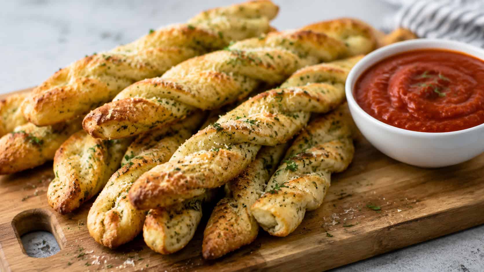 golden Parmesan garlic breadsticks in a wide horizontal shot, twisted sticks with parsley on a board beside a bowl of marinara, natural light, shallow depth of field, landscape orientation