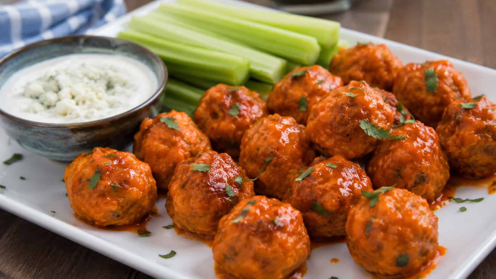 buffalo chicken meatballs in a wide horizontal shot, glossy red-orange meatballs on a platter with a bowl of blue cheese dip and celery, natural light, shallow depth of field, landscape orientation