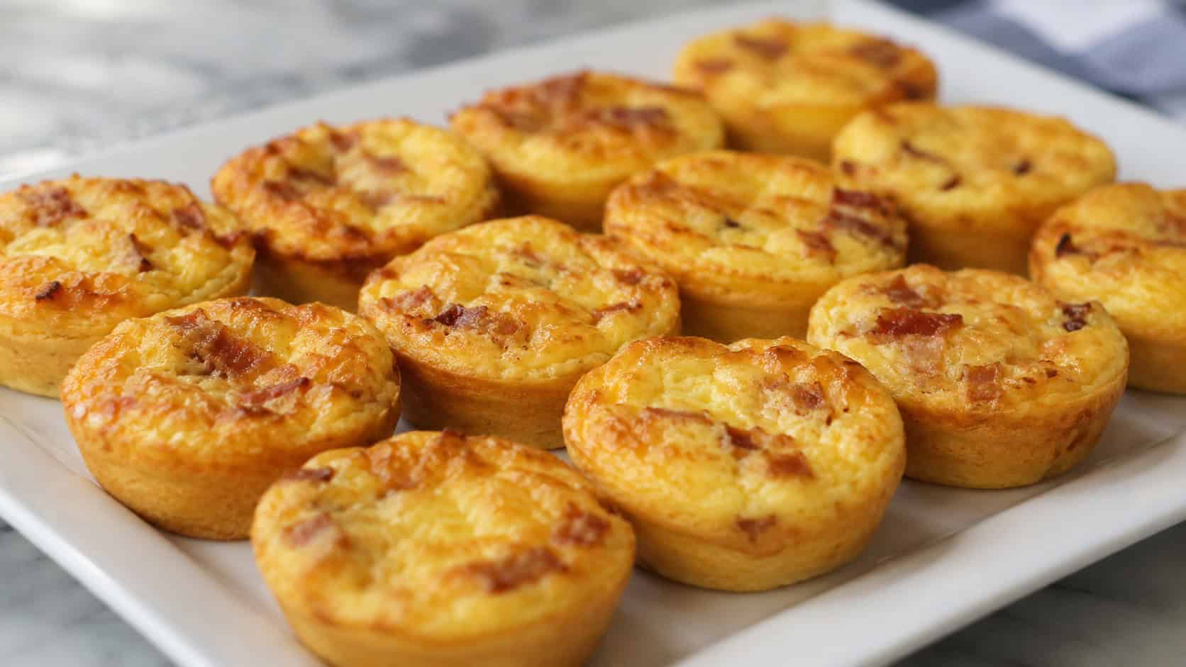 mini quiche Lorraines in a wide horizontal shot, golden individual quiches with flecks of bacon on a white platter, natural light, shallow depth of field, landscape orientation