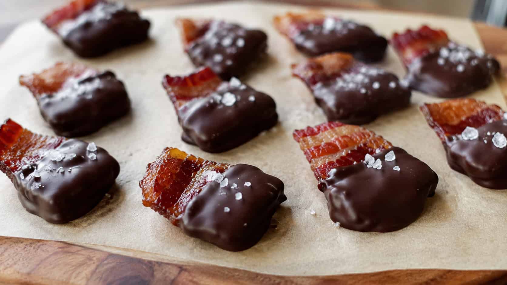 chocolate-covered bacon bites in a wide horizontal shot, half-dipped pieces with flaky salt on parchment-lined board, natural light, shallow depth of field, landscape orientation