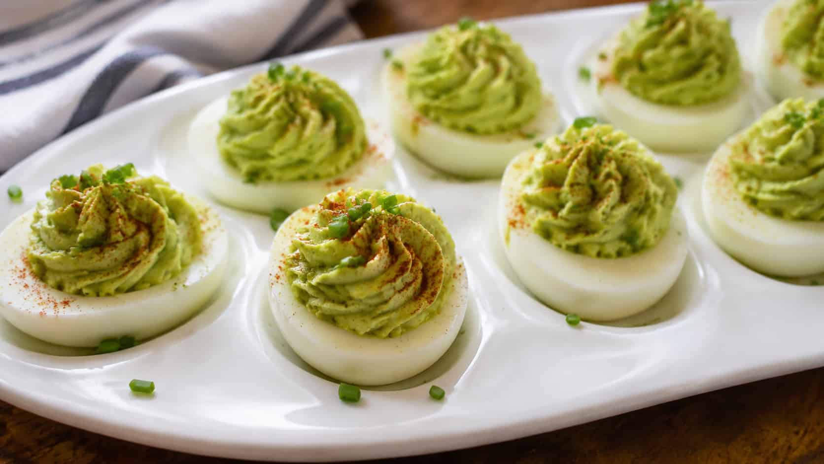 avocado deviled eggs in a wide horizontal shot, pale green piped filling dusted with paprika on a white deviled egg platter, natural light, shallow depth of field, landscape orientation