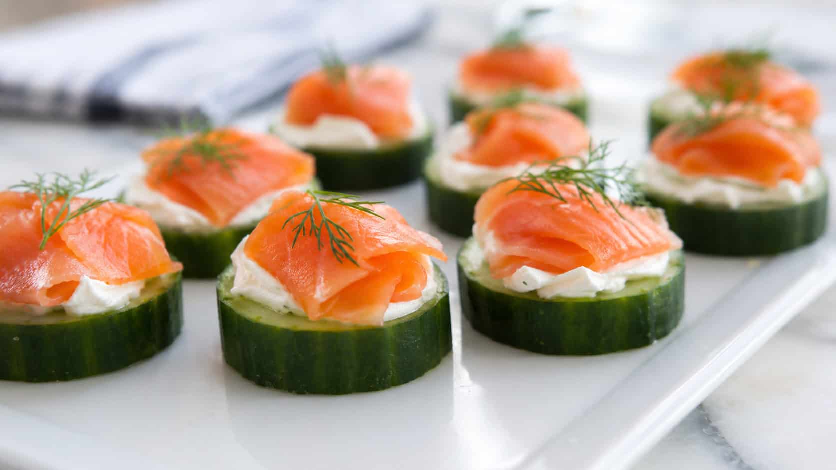 smoked salmon cucumber bites in a wide horizontal shot, neat rounds topped with cream cheese, salmon, and dill on a white serving platter, natural light, shallow depth of field, landscape orientation