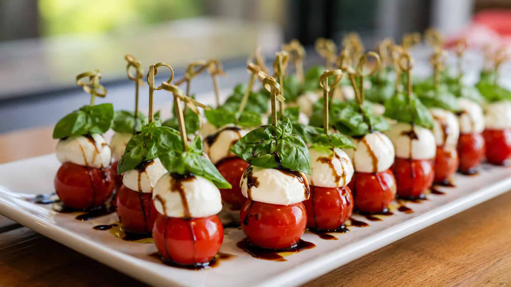 mini caprese skewers in a wide horizontal shot, rows of tomato, mozzarella, and basil on a white platter with a balsamic drizzle, natural light, shallow depth of field, landscape orientation