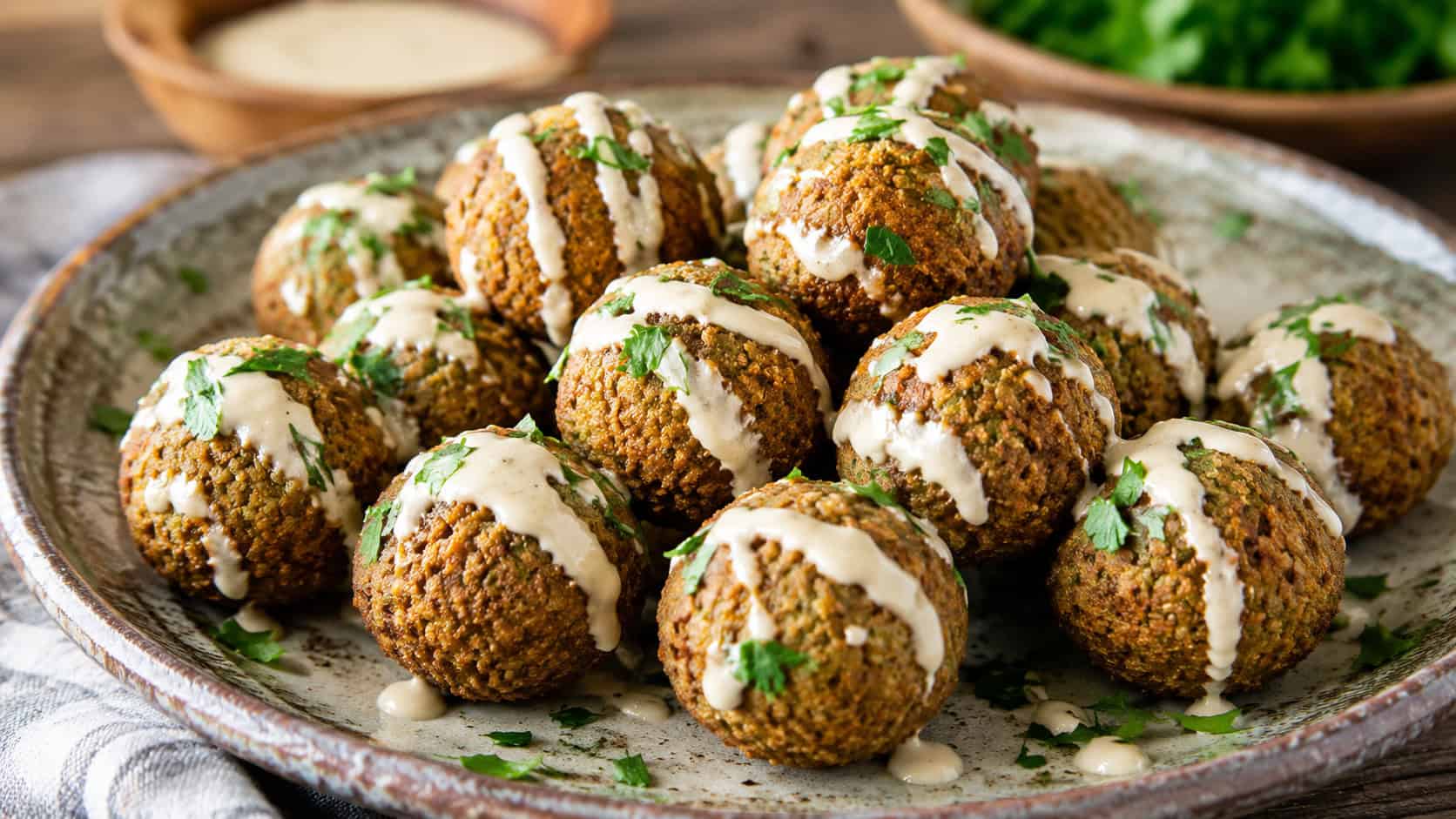mini falafel balls in a wide horizontal shot, golden crisp chickpea balls drizzled with tahini and sprinkled with parsley, on a rustic plate, natural light, shallow depth of field, landscape orientation