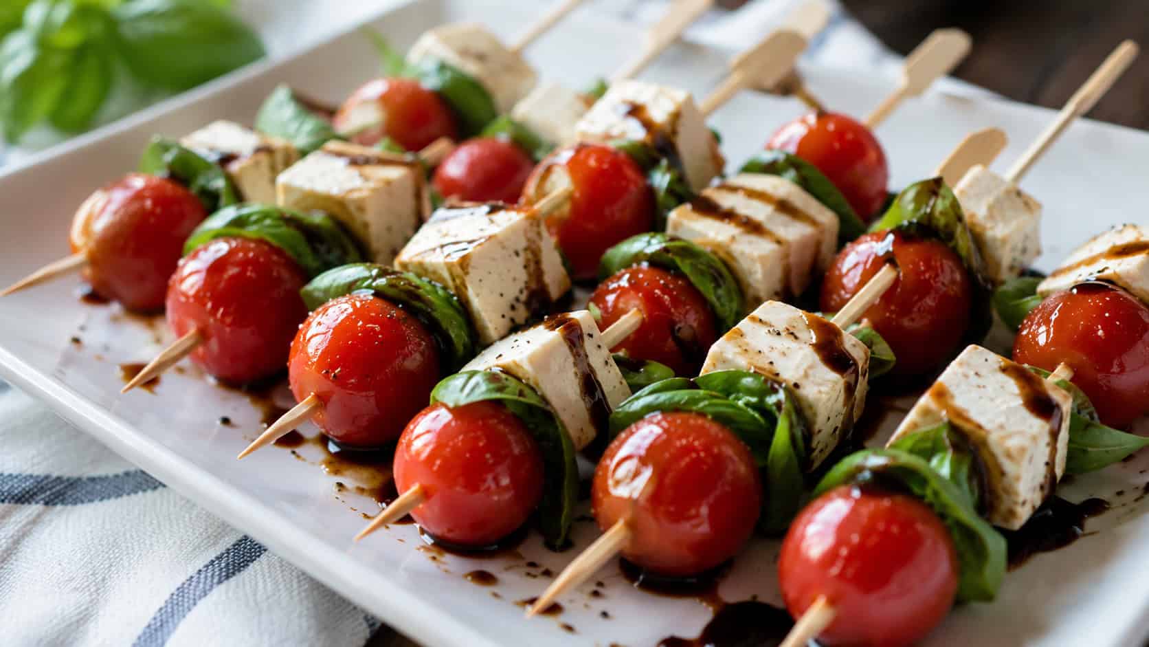 caprese-style skewers in a wide horizontal shot, small wooden picks with red cherry tomatoes, green basil, and white tofu cubes drizzled with balsamic, on a white platter, bright natural light, shallow depth of field, landscape orientation
