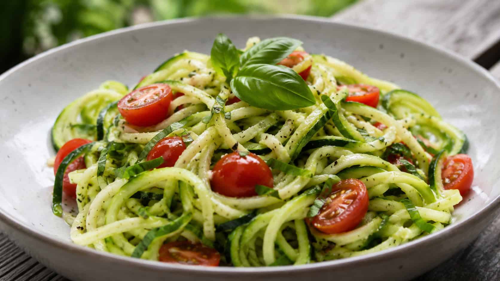 zucchini noodle salad in a wide horizontal shot, pale green spiralized noodles tossed with red cherry tomatoes and basil, served in a shallow bowl, bright natural light, shallow depth of field, landscape orientation