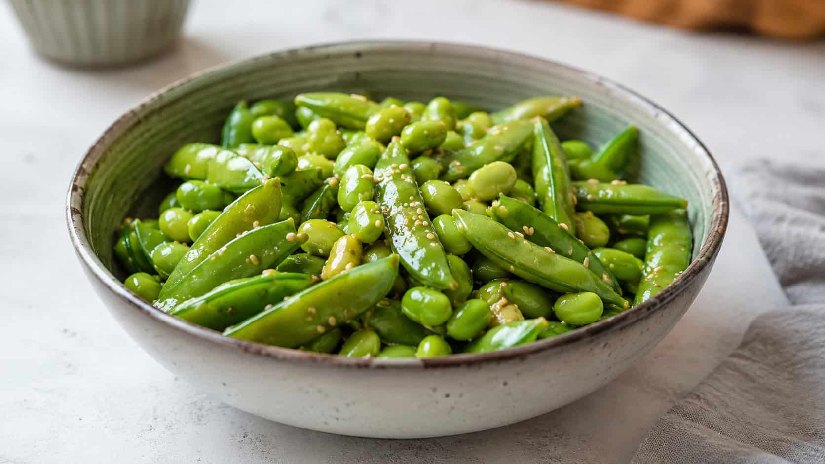an edamame and snap pea bowl in a wide horizontal shot, glossy green pods tossed with sesame seeds in a ceramic bowl, on a light surface, natural light, shallow depth of field, landscape orientation