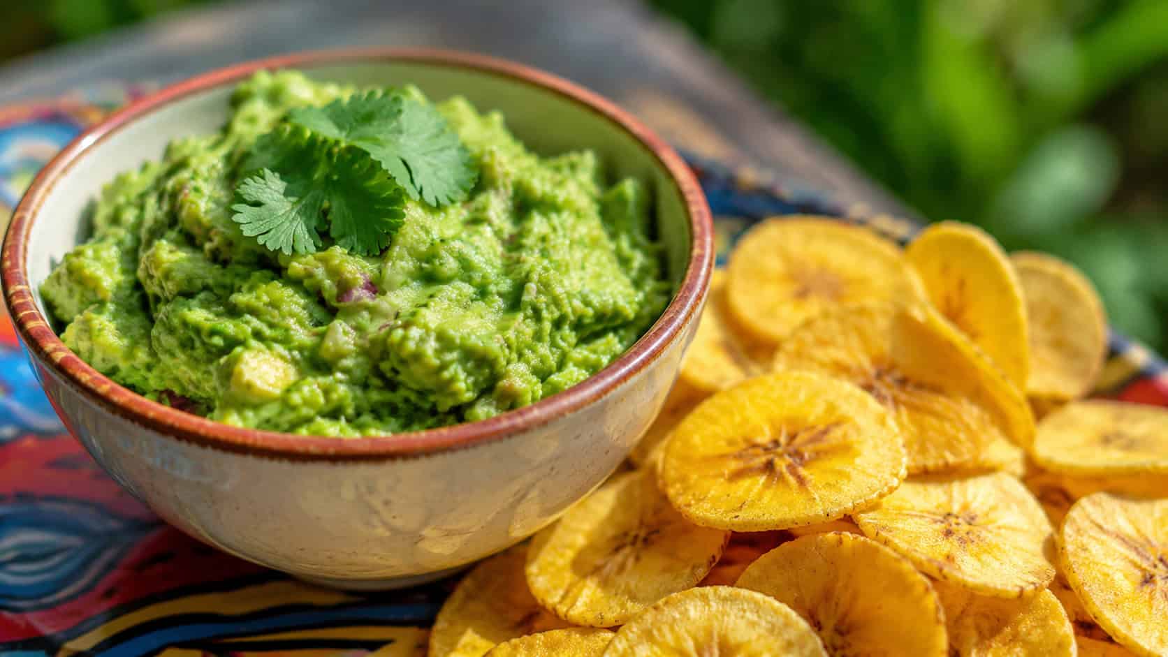guacamole with plantain chips in a wide horizontal shot, a bowl of chunky green guacamole topped with cilantro beside golden plantain chips, on a colorful surface, bright natural light, shallow depth of field, landscape orientation