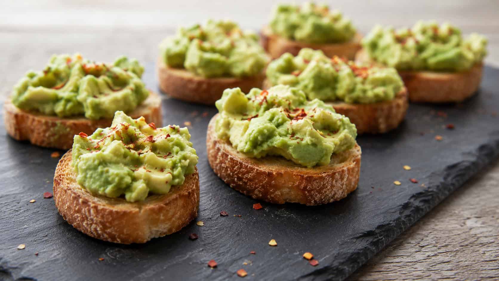 avocado toast bites in a wide horizontal shot, crisp baguette rounds topped with creamy green avocado and red pepper flakes, arranged on a slate board, natural light, shallow depth of field, landscape orientation