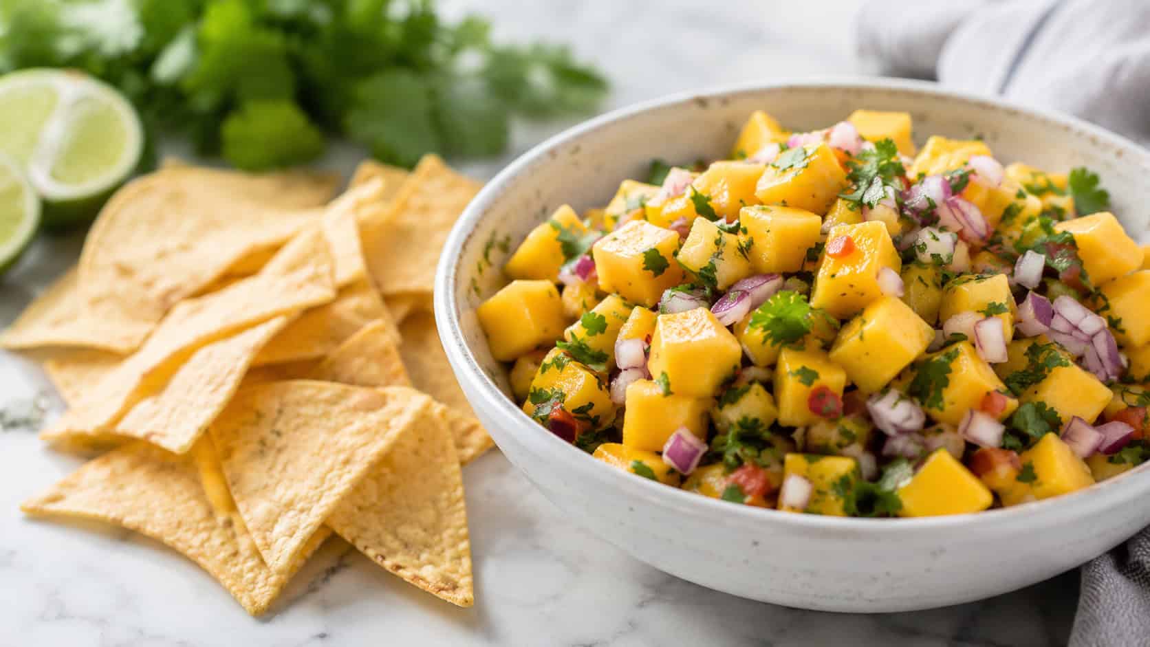 mango salsa in a wide horizontal shot, a vibrant bowl of diced orange mango, red onion, and cilantro beside scattered tortilla chips, on a bright surface, natural light, shallow depth of field, landscape orientation