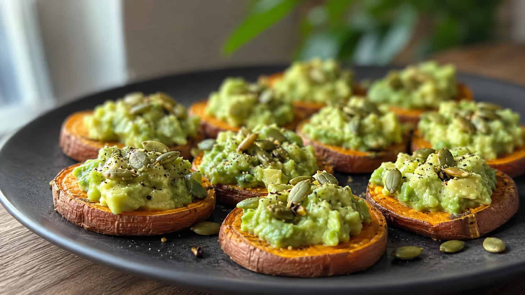sweet potato crostini in a wide horizontal shot, roasted orange sweet potato rounds topped with green avocado and pumpkin seeds, on a dark plate, soft natural light, shallow depth of field, landscape orientation