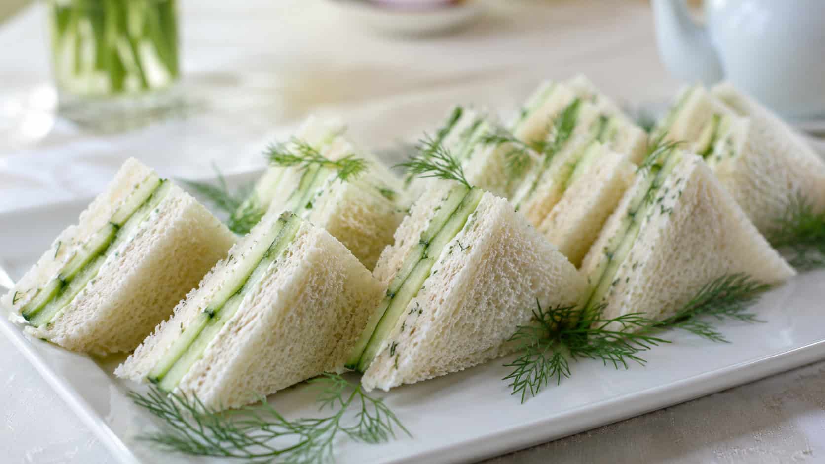 cucumber dill tea sandwiches in a wide horizontal shot, neat crustless triangles layered with cucumber and herb spread, arranged on a white plate with dill sprigs, bright natural light, shallow depth of field, landscape orientation