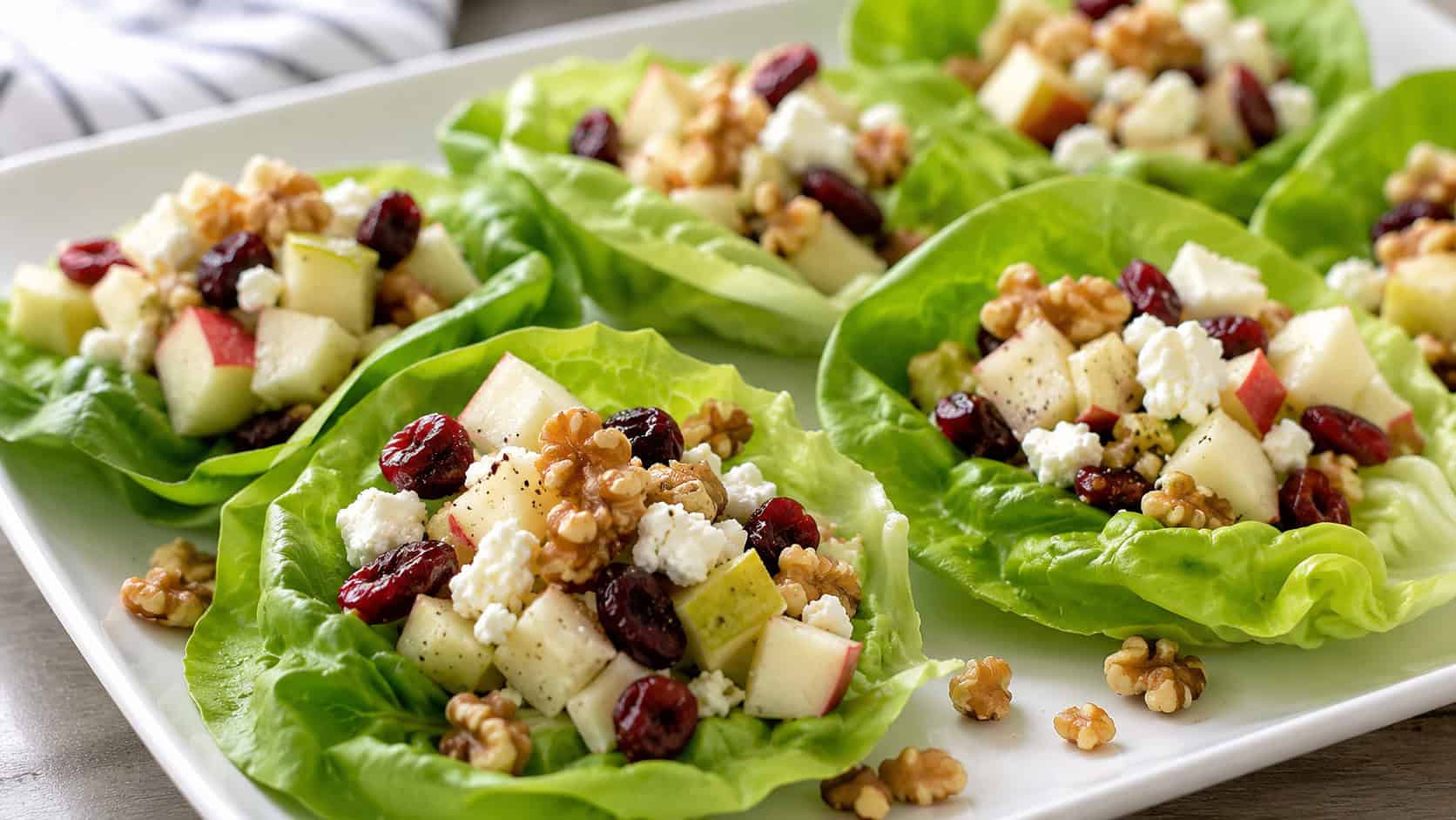 crisp green lettuce cups filled with diced apple, feta, and cranberries in a wide horizontal shot, arranged on a white platter sprinkled with walnuts, natural light, shallow depth of field, landscape orientation
