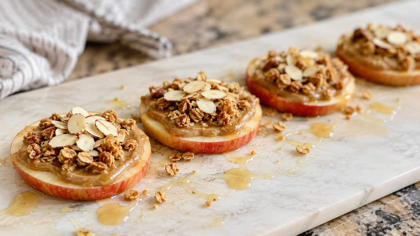 apple rounds topped with almond butter, granola, and sliced almonds in a wide horizontal shot, lined up on a light cutting board drizzled with honey, natural light, shallow depth of field, landscape orientation