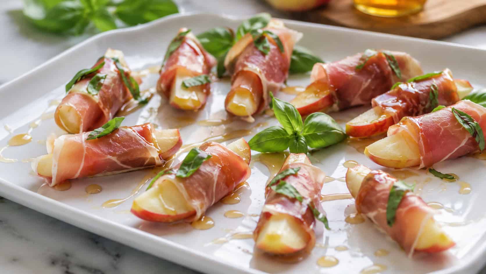 apple wedges wrapped in thin prosciutto with basil in a wide horizontal shot, arranged on a white platter drizzled with honey, natural light, shallow depth of field, landscape orientation
