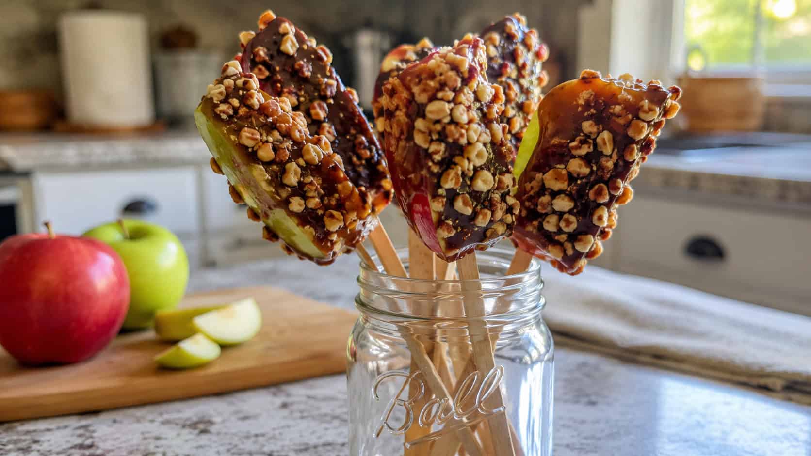 apple slices on sticks glazed with glossy maple and rolled in pecans and chocolate in a wide horizontal shot, standing in a jar on a kitchen counter, natural light, shallow depth of field, landscape orientation