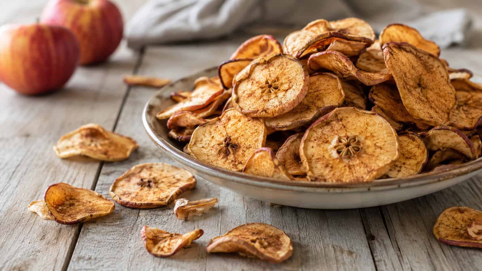thin crispy cinnamon apple chips in a wide horizontal shot, piled in a shallow bowl and scattered across a light wooden table, natural light, shallow depth of field, landscape orientation