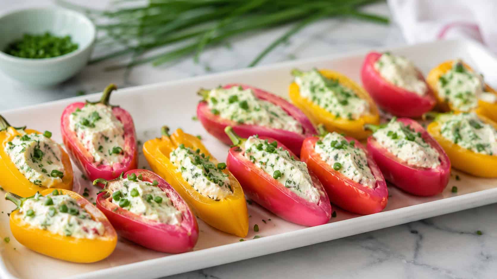 stuffed mini peppers in pink and yellow arranged on a white platter in a wide horizontal shot, creamy filling, fresh chives, bright cheerful natural light, shallow depth of field, landscape orientation