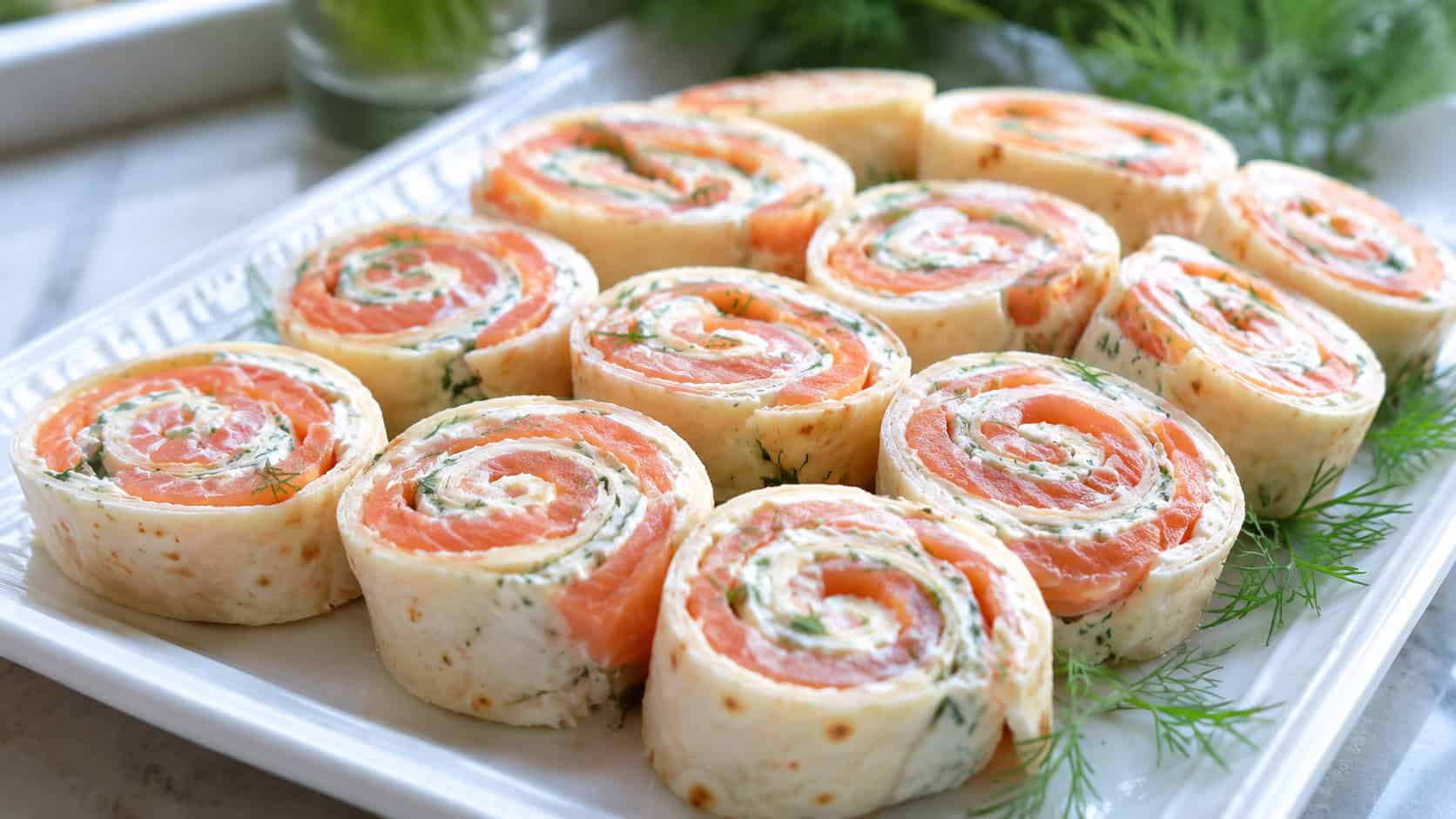 salmon and cream cheese pinwheels arranged cut-side up on a white platter in a wide horizontal shot, pink swirls, fresh dill, bright natural light, shallow depth of field, landscape orientation
