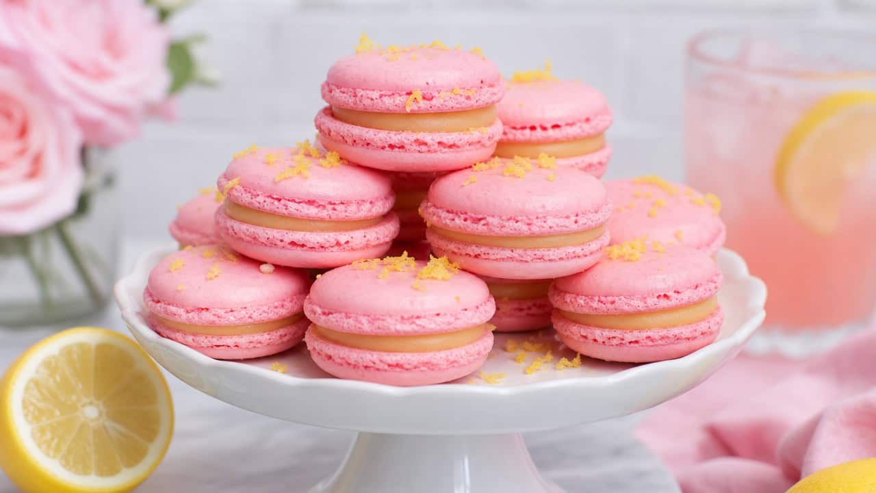 pink lemonade macarons stacked on a white cake stand in a wide horizontal shot, smooth glossy shells, lemon zest accents, soft pastel styling, bright natural light, shallow depth of field, landscape orientation