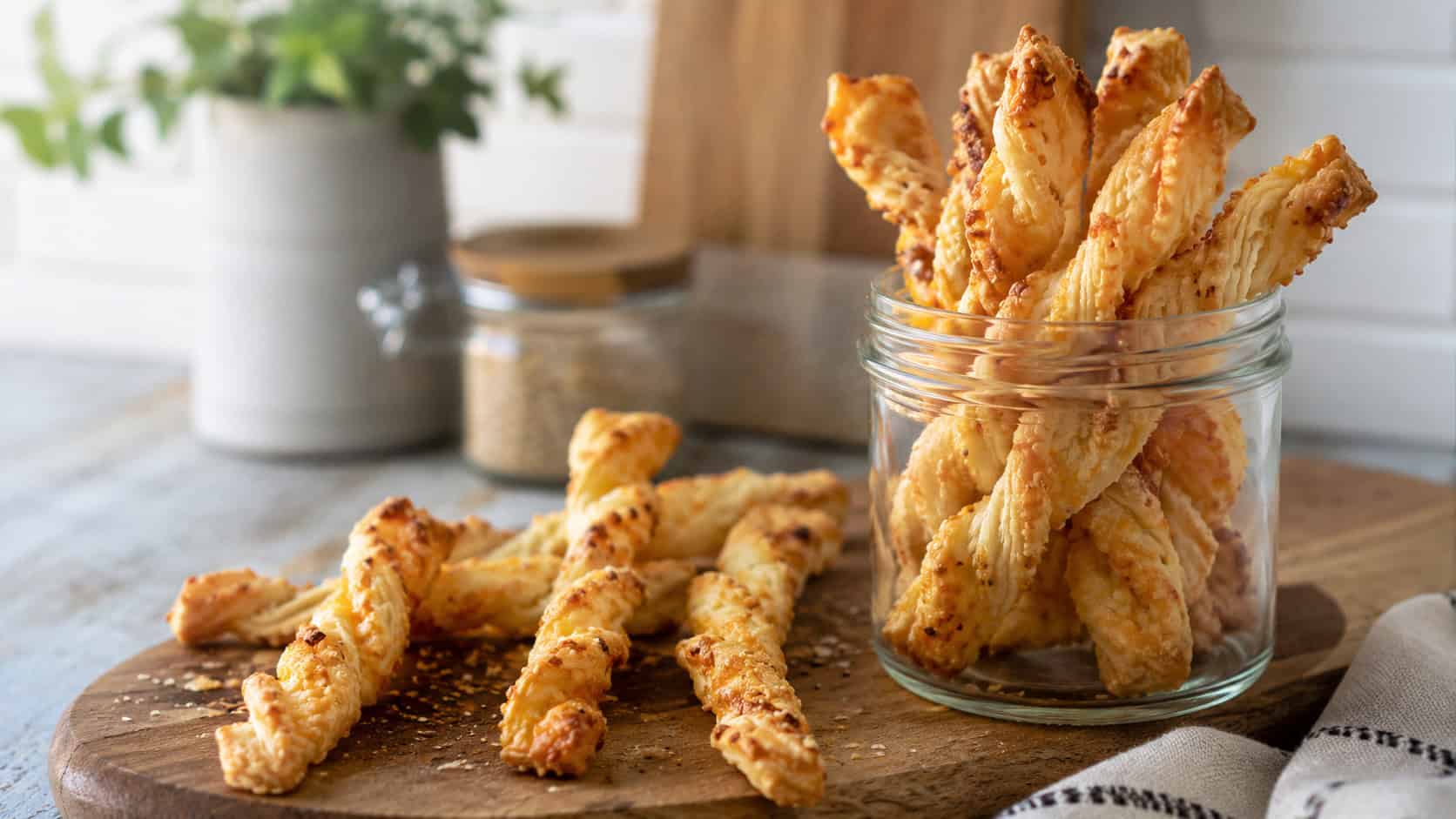 golden savory puff pastry twists standing in a jar and scattered on a board in a wide horizontal shot, flaky cheesy layers, warm natural light, shallow depth of field, landscape orientation