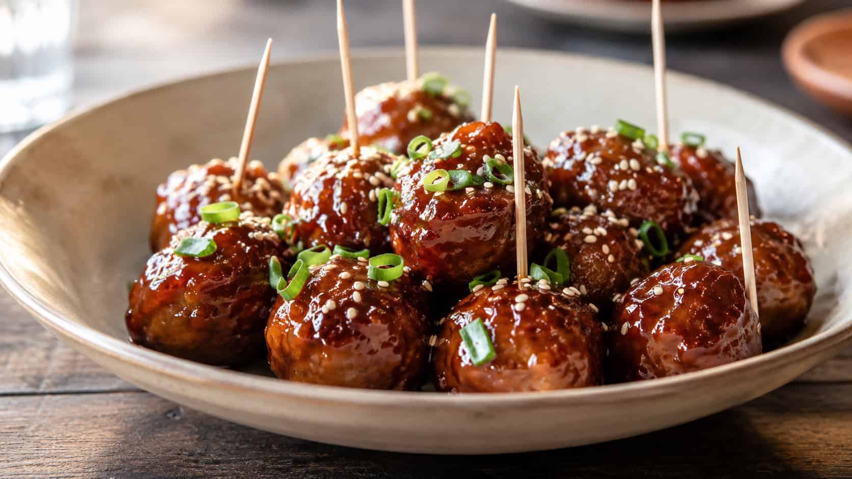 glossy ginger teriyaki meatballs in a wide shallow bowl in a horizontal shot, sticky glaze, sesame seeds, sliced green onions, toothpicks ready, warm natural light, shallow depth of field, landscape orientation
