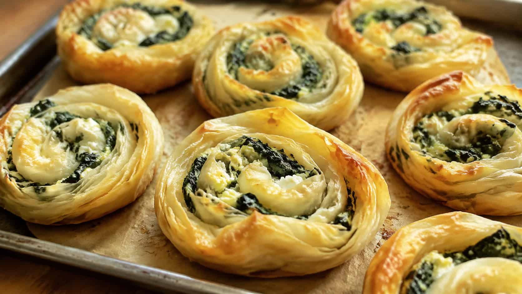 golden spinach and feta pastry swirls on a baking sheet in a wide horizontal shot, flaky layers, green and white filling visible, warm natural light, shallow depth of field, landscape orientation