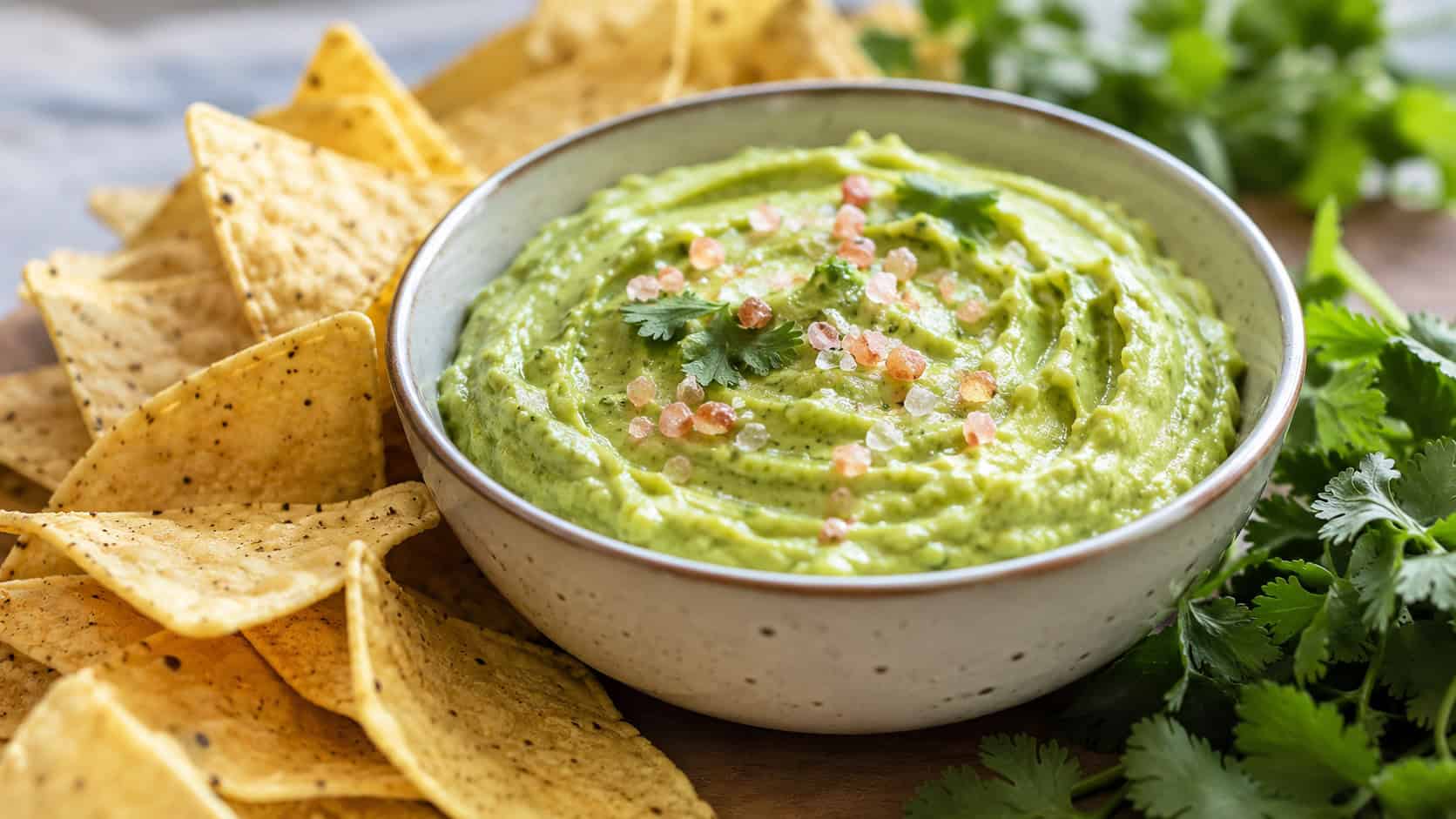 creamy avocado dip in a bowl in a wide horizontal shot, sprinkle of pink salt on top, tortilla chips fanned around, fresh cilantro, bright natural light, shallow depth of field, landscape orientation