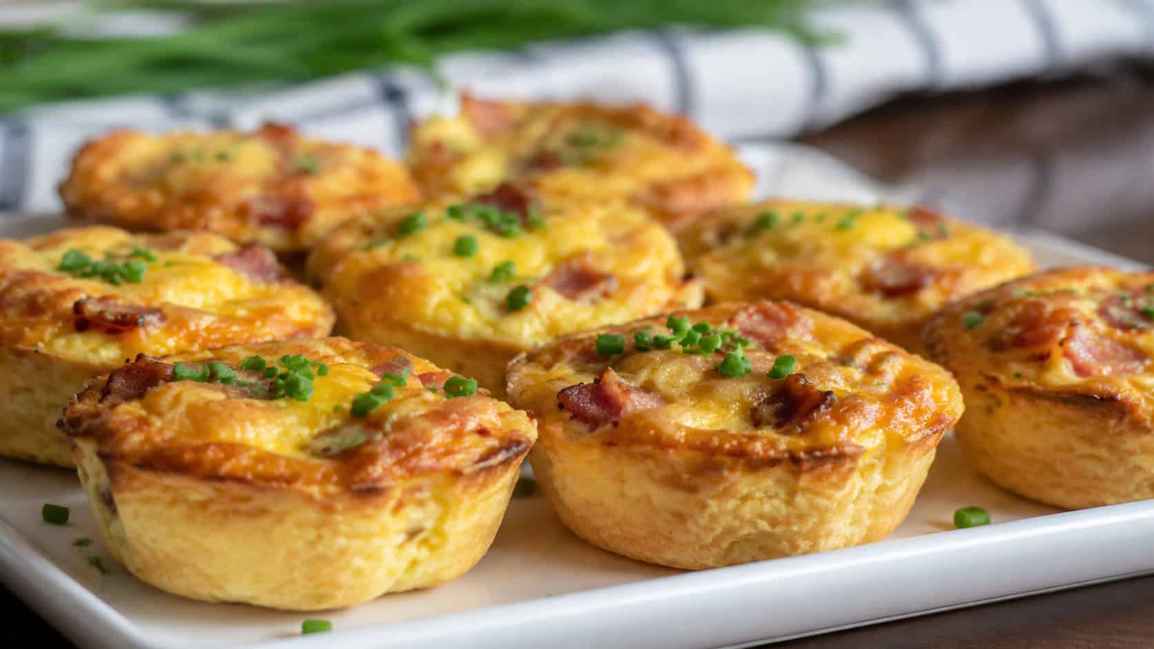 golden mini quiches with bacon and melted cheese in a wide horizontal shot, arranged on a serving platter with chives, natural light, shallow depth of field, landscape orientation