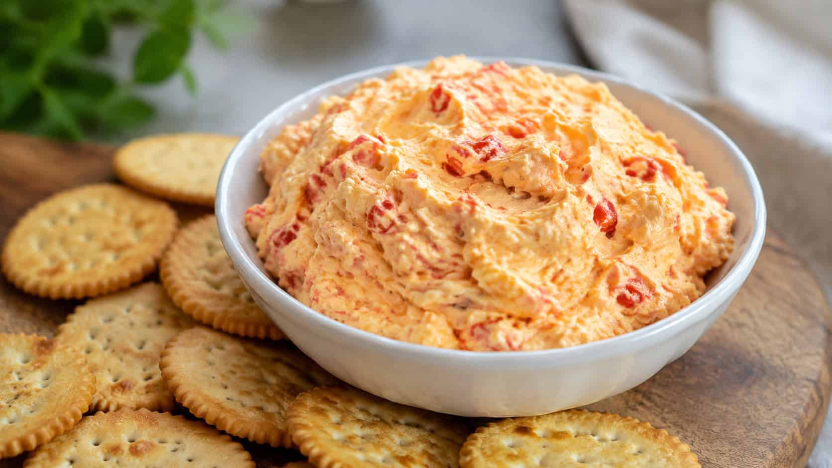 a bowl of creamy pimento cheese spread flecked with red pimentos in a wide horizontal shot, surrounded by crackers on a board, natural light, shallow depth of field, landscape orientation