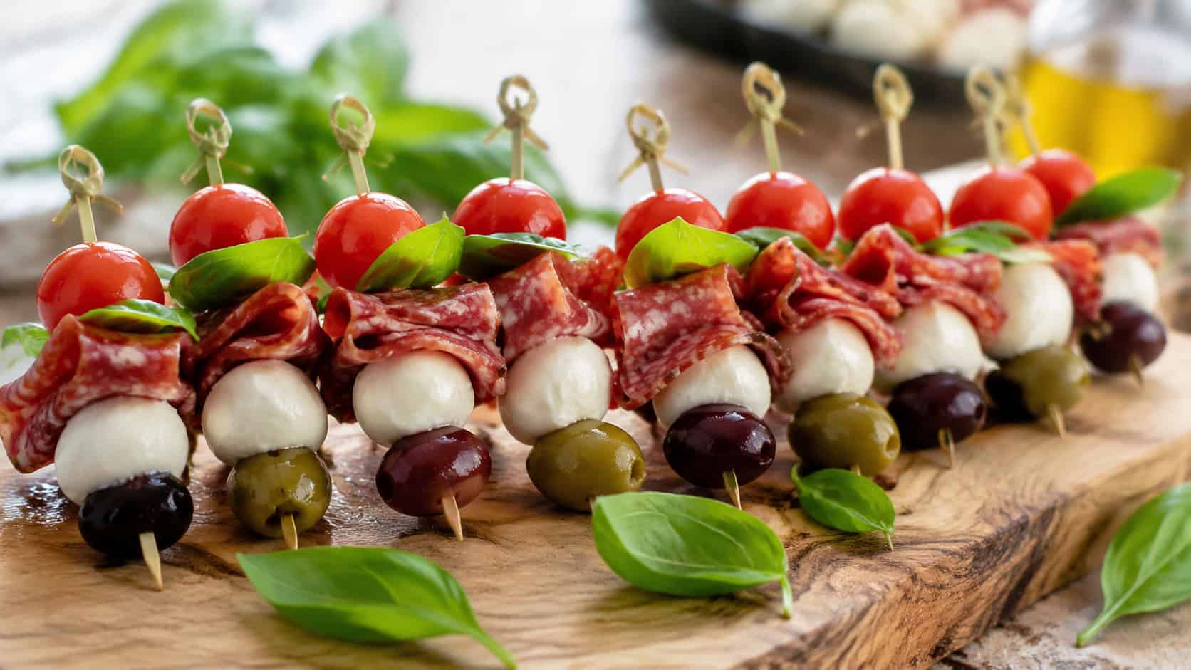 colorful skewers of folded salami, mozzarella, olives, and cherry tomatoes in a wide horizontal shot, lined up on a wooden board with basil, natural light, shallow depth of field, landscape orientation