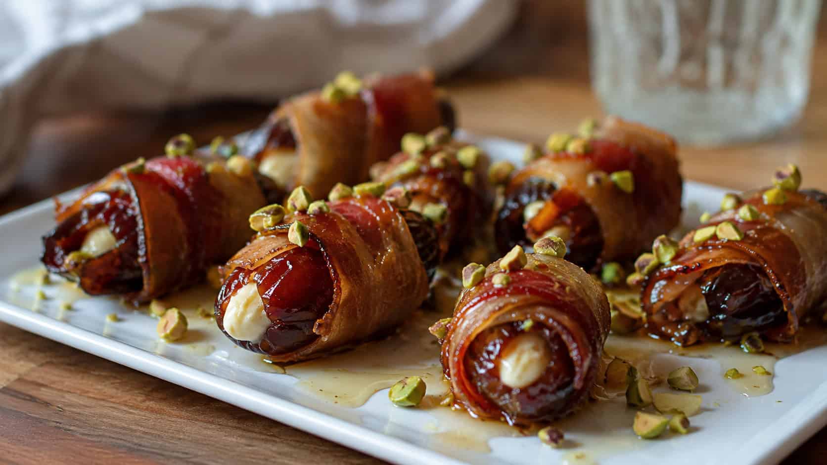 bacon-wrapped stuffed dates topped with chopped pistachios and honey in a wide horizontal shot, on a small serving plate, natural light, shallow depth of field, landscape orientation
