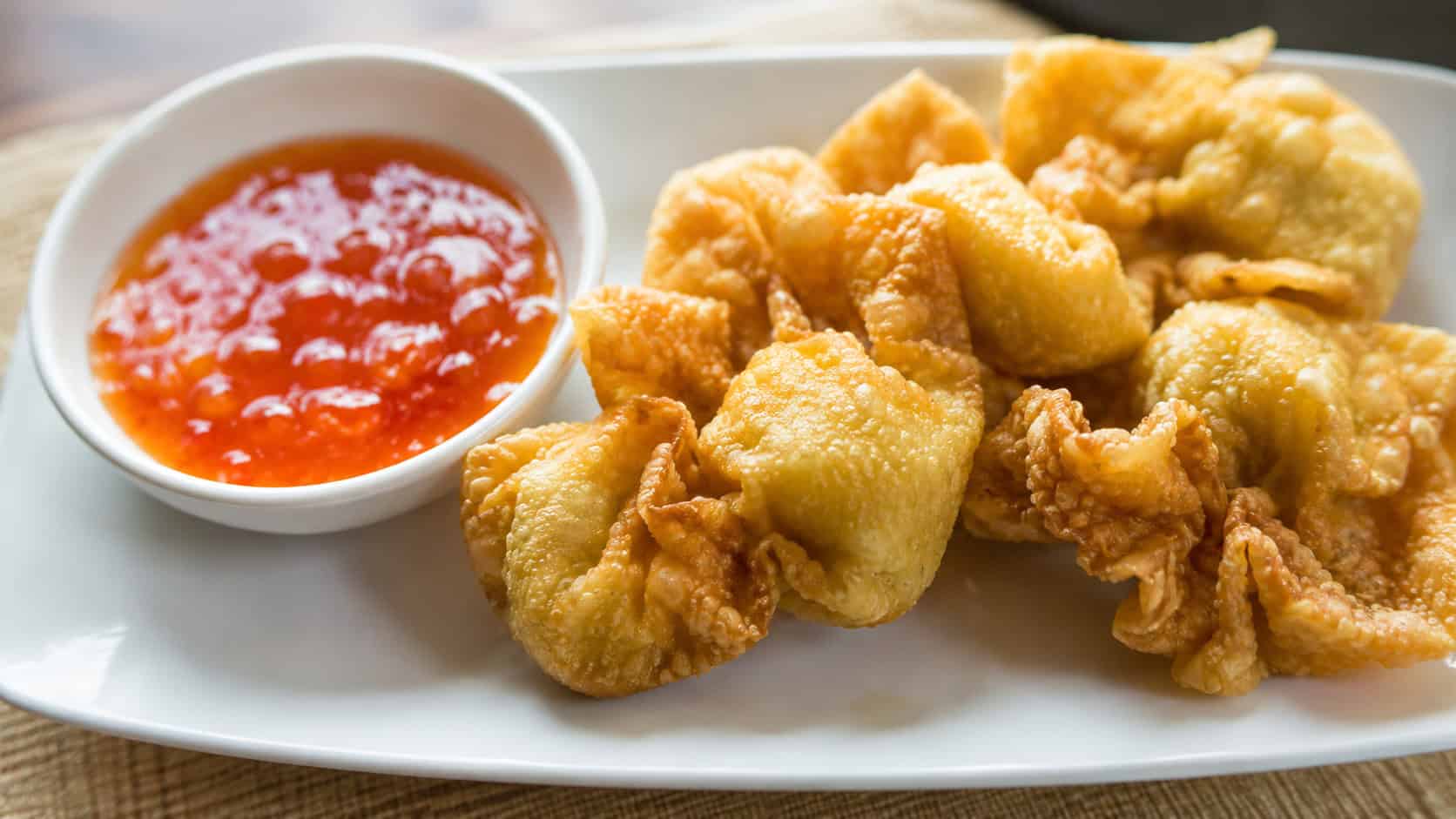 sweet and sour pork wontons in a wide horizontal shot, golden parcels beside a bowl of glossy red-orange dipping sauce on a white plate, natural light, shallow depth of field, landscape orientation