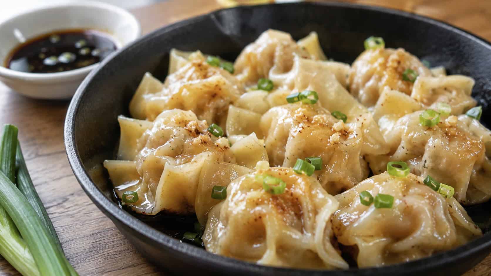 ginger garlic shrimp wontons in a wide horizontal shot, glistening pan-steamed parcels in a black skillet with scallions and a small soy dish, natural light, shallow depth of field, landscape orientation