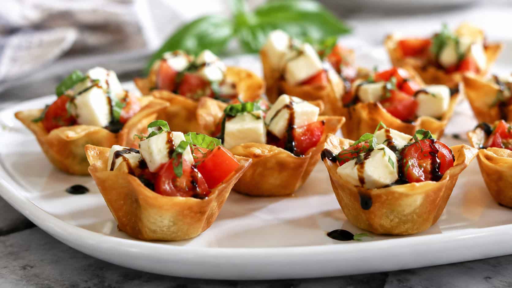 caprese wonton bites in a wide horizontal shot, crisp cups holding red tomato and white mozzarella with green basil and balsamic drizzle on a white platter, bright natural light, shallow depth of field, landscape orientation