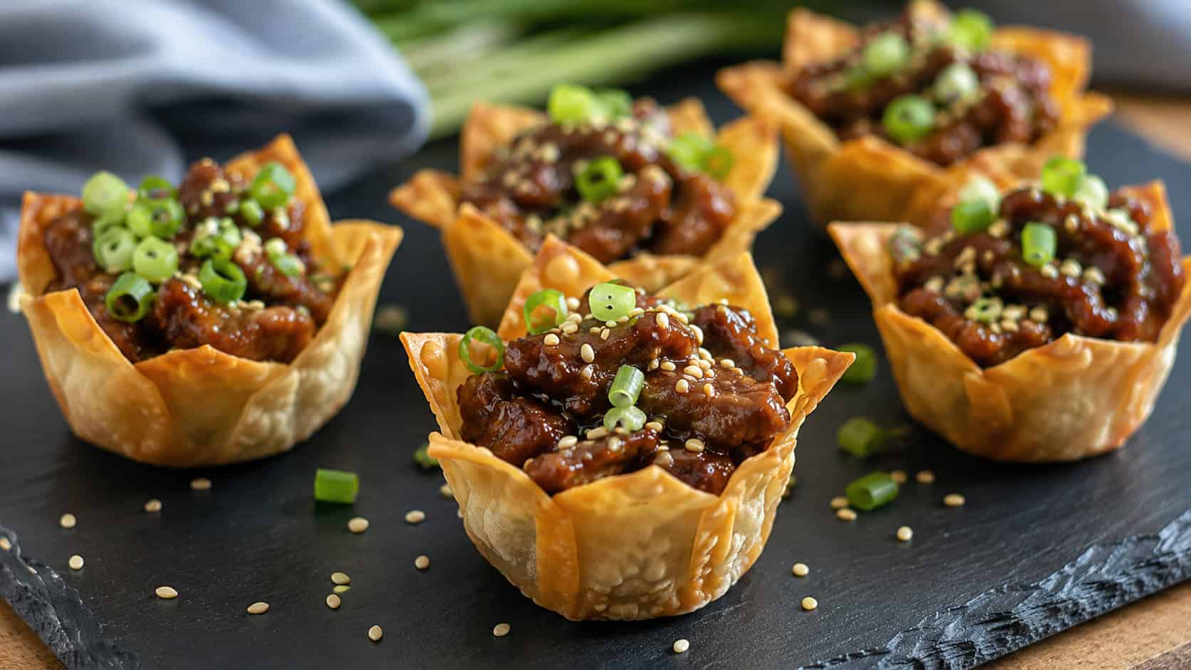 teriyaki beef wonton cups in a wide horizontal shot, glossy beef in golden shells topped with sesame seeds and scallions on a dark slate board, natural light, shallow depth of field, landscape orientation