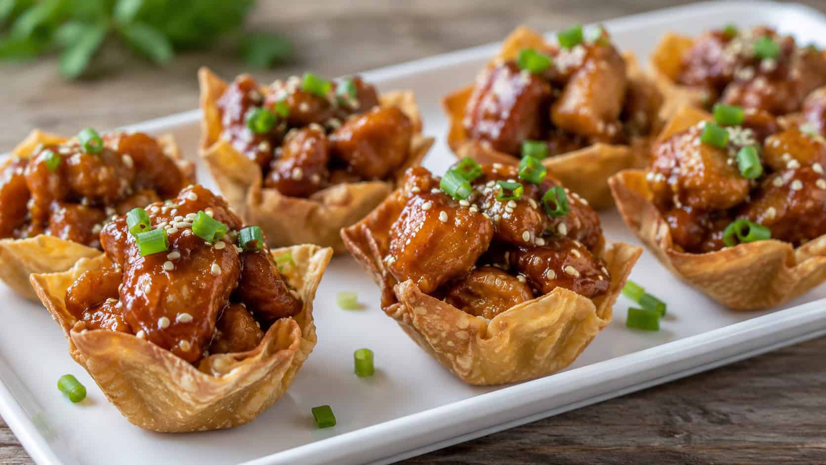 sesame chicken wonton cups in a wide horizontal shot, glossy chicken in golden shells topped with sesame seeds and scallions on a white platter, natural light, shallow depth of field, landscape orientation
