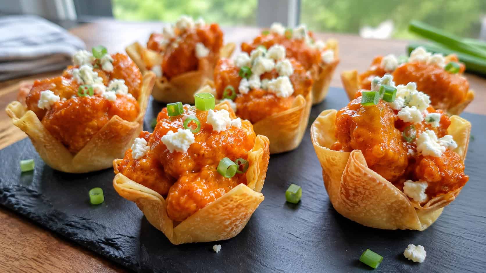 buffalo cauliflower wonton cups in a wide horizontal shot, orange-glazed cauliflower in crisp shells topped with white feta on a slate board, natural light, shallow depth of field, landscape orientation