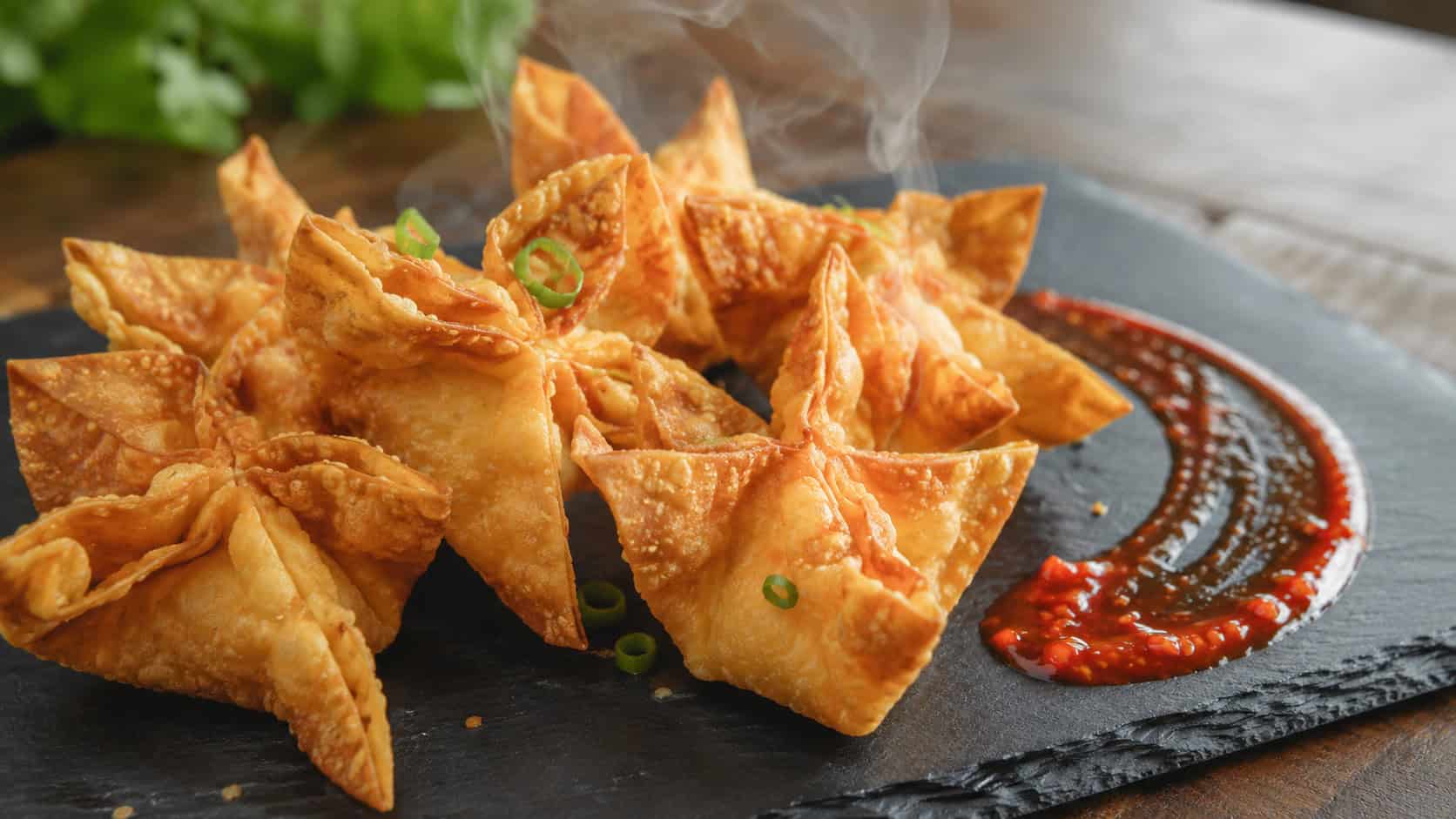 crispy fried shrimp rangoon in a wide horizontal shot, piled on a dark slate board with a swoosh of red chili sauce, steam rising, natural light, shallow depth of field, landscape orientation