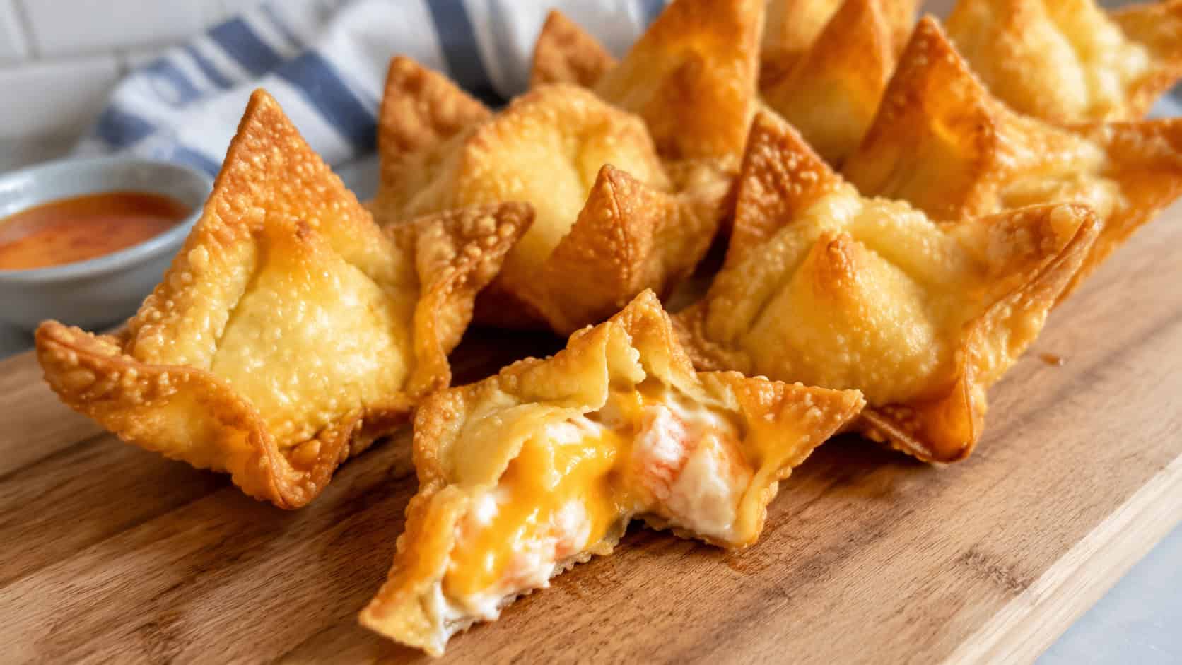 golden cheesy crab wontons in a wide horizontal shot, fanned across a wooden cutting board with one broken open to show melty filling, warm natural light, shallow depth of field, landscape orientation
