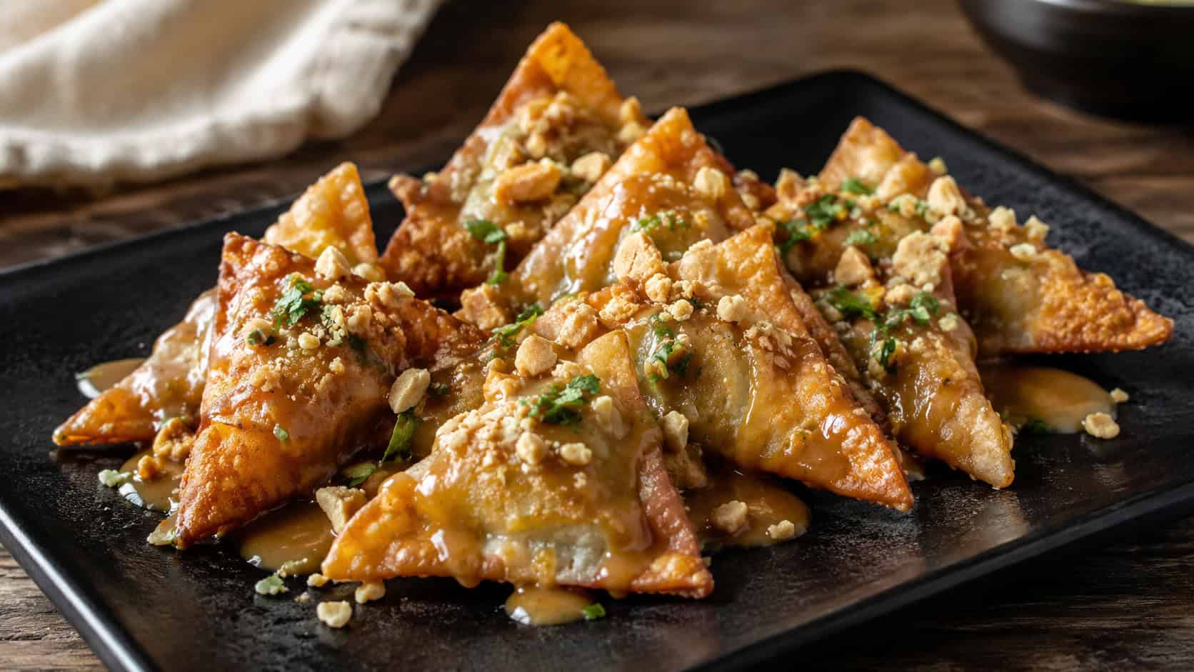 Thai peanut chicken wontons in a wide horizontal shot, crispy triangles on a dark plate drizzled with peanut sauce and chopped peanuts, natural light, shallow depth of field, landscape orientation
